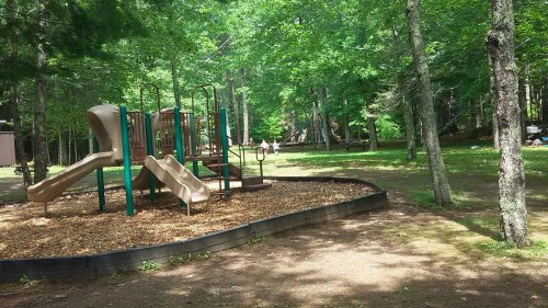 A playground with slides surrounded by trees, set in a grassy area with children playing in the background.