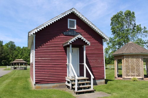 A red wooden building with white trim and steps, surrounded by green grass and trees, with a gazebo nearby.