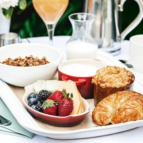 A breakfast tray featuring yogurt, granola, fresh fruit, a muffin, and a croissant, with a drink in the background.