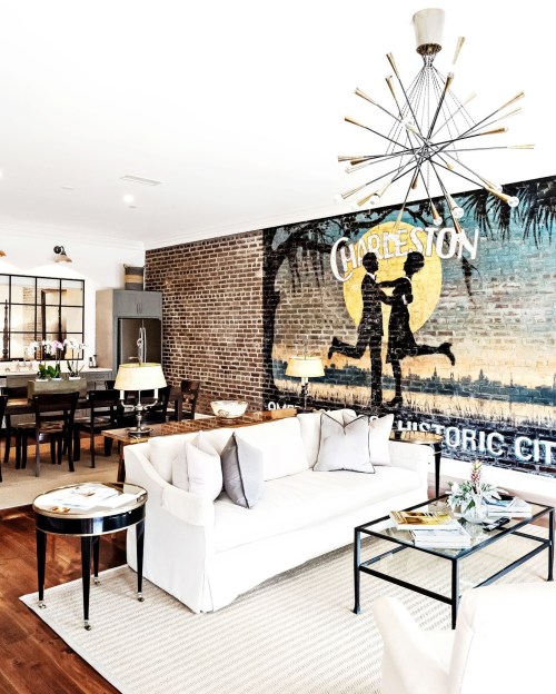 Bright living room with a white sofa, coffee table, and a mural of Charleston on a brick wall. Modern lighting fixtures.