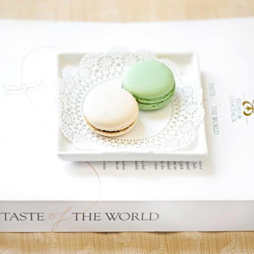 Two macarons, one white and one green, on a decorative plate atop a book titled "Taste of the World."