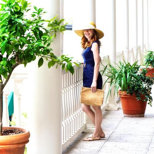 A woman in a navy dress and wide-brimmed hat stands by a balcony, holding a clutch, surrounded by potted plants.