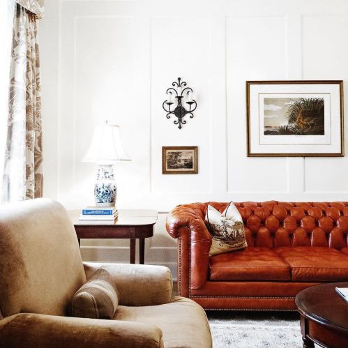 Cozy living room featuring a brown leather couch, armchair, lamp, and framed artwork on white walls.