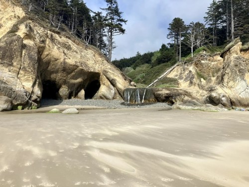 A sandy beach with rocky cliffs, a small waterfall, and lush trees in the background under a cloudy sky.