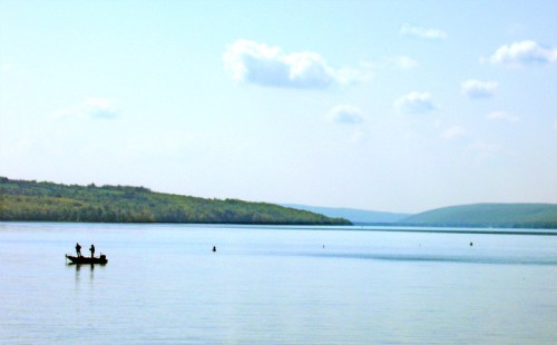 A serene lake scene with a small boat and two people fishing, surrounded by lush green hills under a blue sky.