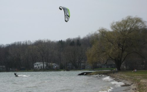 A kite surfer glides over water with a large kite above, while a tree and houses line the shore in the background.