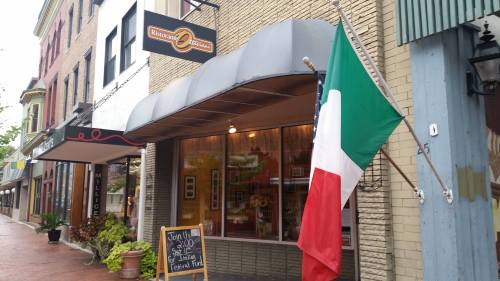 A street view of a restaurant with an Italian flag outside, surrounded by shops on a rainy day.