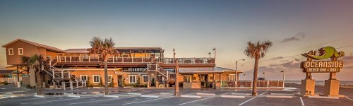 A beachside bar and grill with palm trees, outdoor seating, and a sign reading "Oceanside" at sunset.