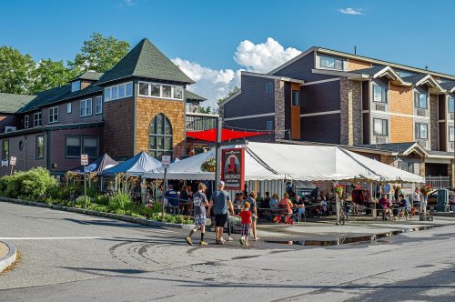 A bustling outdoor dining area with a large tent, people walking, and buildings in the background under a blue sky.