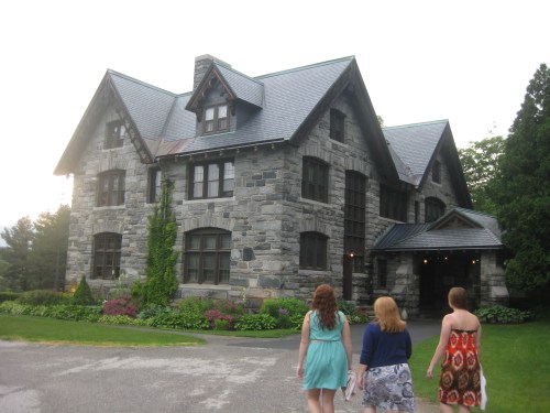 Three women walk towards a large stone house surrounded by greenery and colorful flowers on a sunny day.