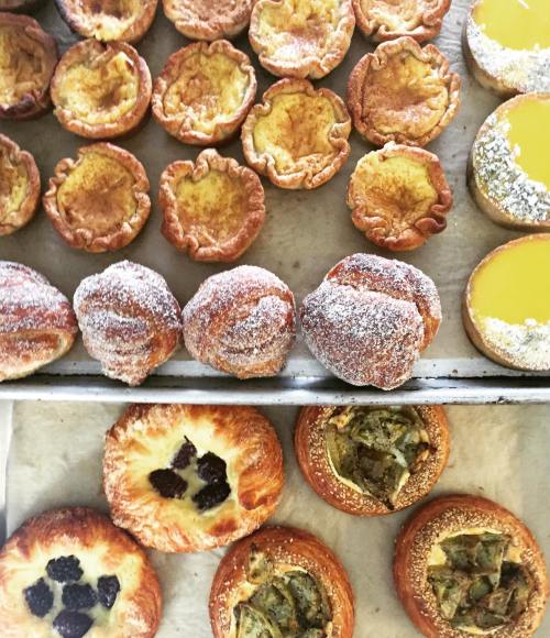 A variety of baked pastries, including custard tarts, powdered sugar doughnuts, and fruit-filled danishes.
