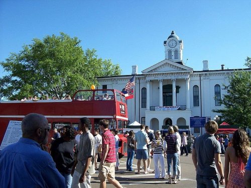 A bustling outdoor event with people gathered near a red double-decker bus and a historic building in the background.