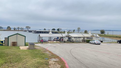 A lakeside view featuring a parking area, a building by the water, and cloudy skies in the background.