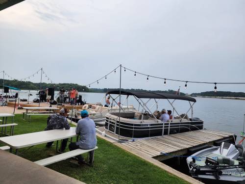 A lakeside scene with people enjoying a performance, boats docked, and string lights overhead.