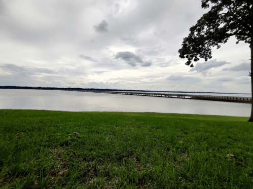 A calm lake scene with a grassy shore, a distant pier, and overcast skies.