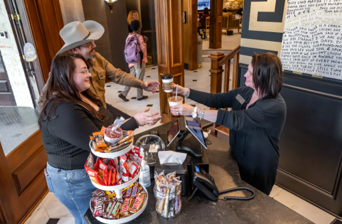 A woman at a reception desk hands drinks to a man and woman, with snacks displayed on a nearby stand.