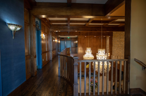 A wooden staircase leads to a spacious hallway with chandeliers and elegant wall sconces in a luxurious interior.