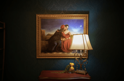 A framed painting of a woman in a red dress, beside a lamp on a wooden table against a dark blue wall.