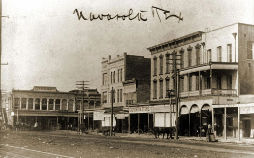 Historic black-and-white photo of a street in New Orleans, featuring buildings, storefronts, and horse-drawn carriages.