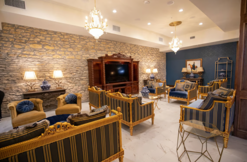Elegant lounge with stone walls, ornate furniture, and chandeliers, featuring blue and gold decor and a large TV.