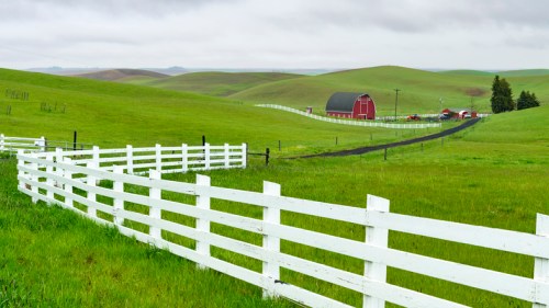 A lush green landscape with rolling hills, a red barn, and a white fence under a cloudy sky.