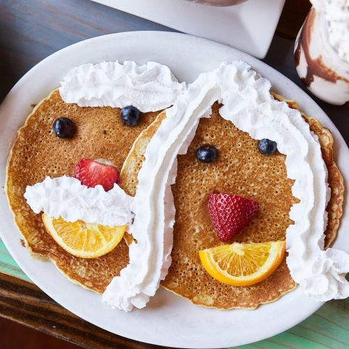 Two pancakes decorated to look like bunny faces, with whipped cream, fruit, and blueberries for eyes and noses.