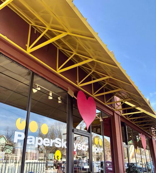 A storefront with large windows and a yellow awning, featuring a pink heart decoration on the wall.