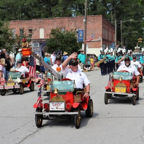 A parade scene with people in small fire trucks waving, surrounded by spectators and marching bands.