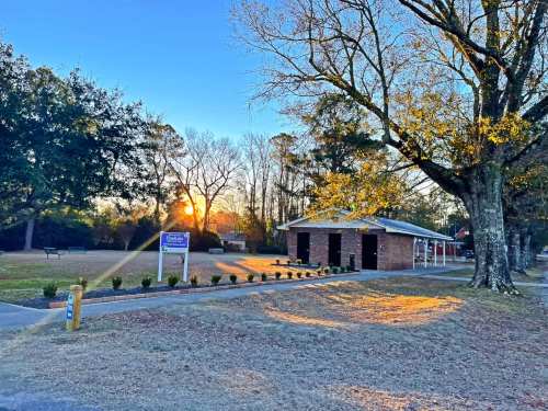 A brick building in a park at sunset, surrounded by trees and a grassy area with a few benches.