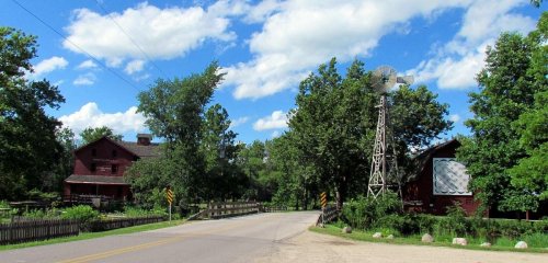 A scenic rural road with a red barn, windmill, and lush green trees under a bright blue sky with fluffy clouds.