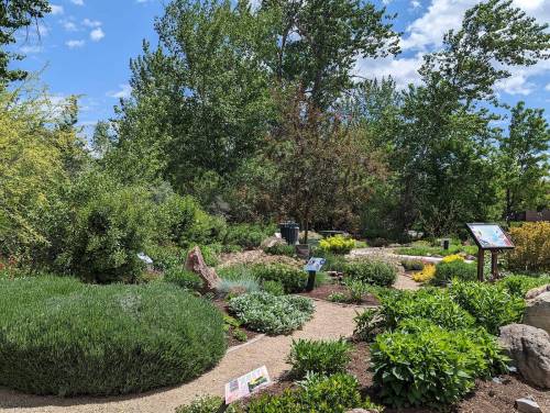 A lush garden with various plants, pathways, and informational signs under a blue sky with scattered clouds.