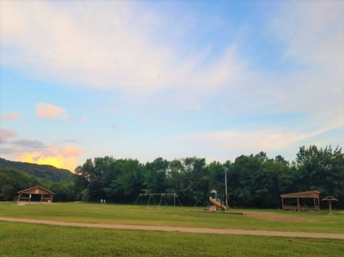A playground with swings and a slide, surrounded by trees, under a colorful sky at dusk.