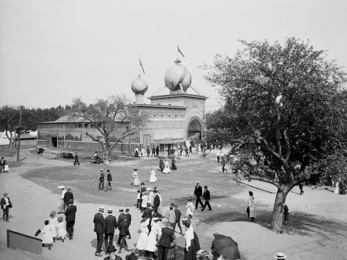 Historic black-and-white photo of a fairground with a large building, crowds of people, and trees in the background.