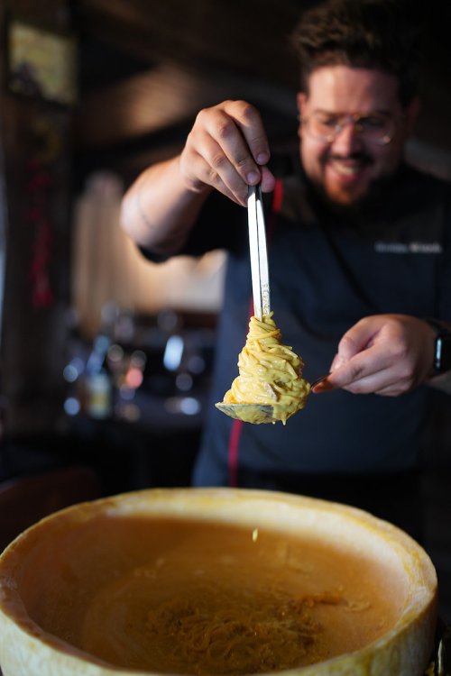 A chef uses tongs to lift pasta from a large cheese wheel, smiling in a cozy restaurant setting.