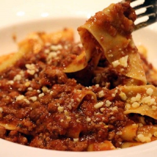 A fork lifting a portion of pappardelle pasta topped with rich meat sauce and sprinkled with cheese.