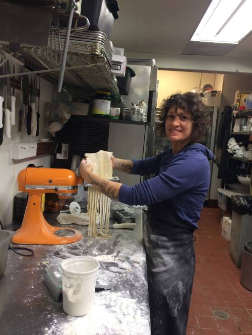 A person with curly hair smiles while holding freshly made pasta in a kitchen, with cooking tools and ingredients around.