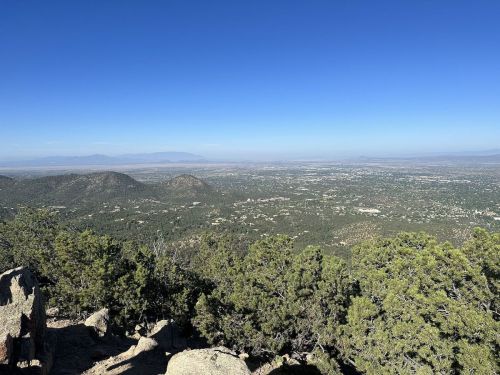 A panoramic view of a valley surrounded by mountains, with clear blue skies and scattered greenery in the foreground.