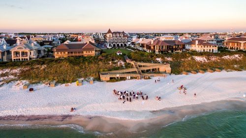 Aerial view of a beach with white sand, houses, and people gathered near the water at sunset.