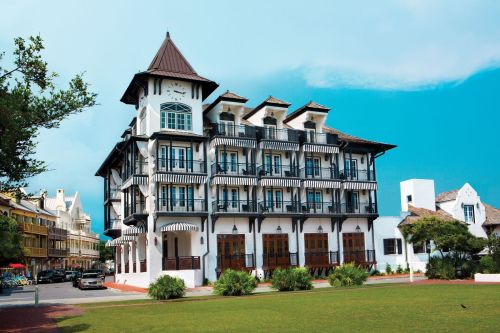 A charming white building with a clock tower, featuring balconies and a lush green lawn in front under a blue sky.