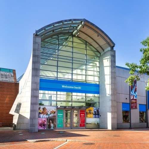 Modern building entrance with a glass roof and a "Welcome back!" sign, surrounded by brick pavement and greenery.