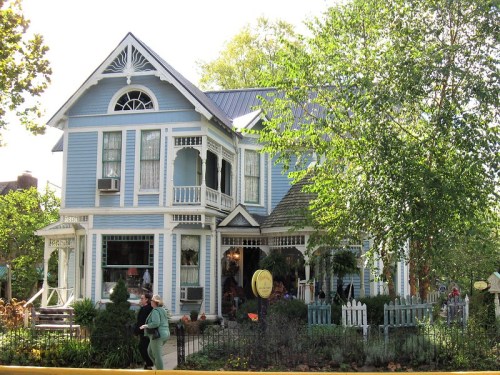 A charming blue Victorian house with intricate details, surrounded by greenery and a picket fence.