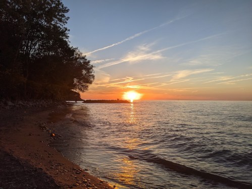 A serene sunset over a calm lake, with a sandy shore and trees silhouetted against the colorful sky.