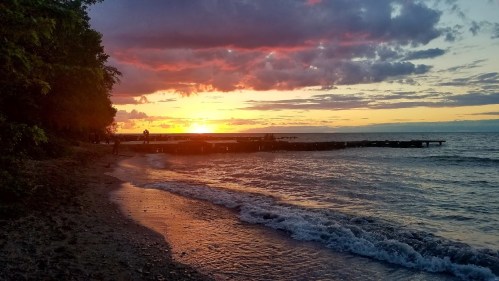 A serene beach at sunset, with colorful clouds reflecting on the water and a pier extending into the lake.