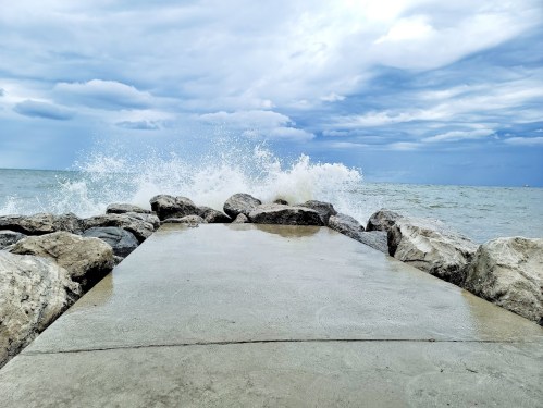 A rocky shoreline with waves crashing against a concrete path under a cloudy sky.
