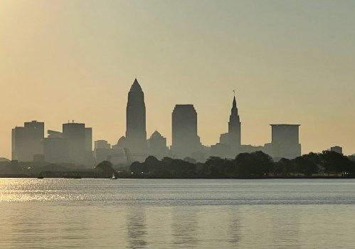 Silhouette of a city skyline at sunrise, with tall buildings reflected in calm water.