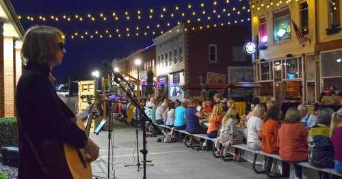 A musician performs on a street filled with people enjoying an outdoor gathering under string lights at night.