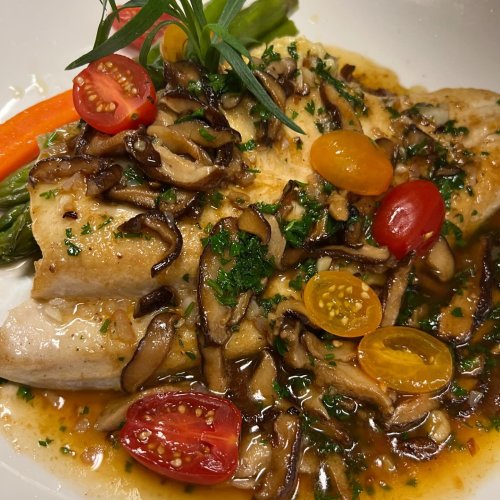 A plate of fish topped with sautéed mushrooms, cherry tomatoes, and herbs, served with asparagus and a colorful garnish.