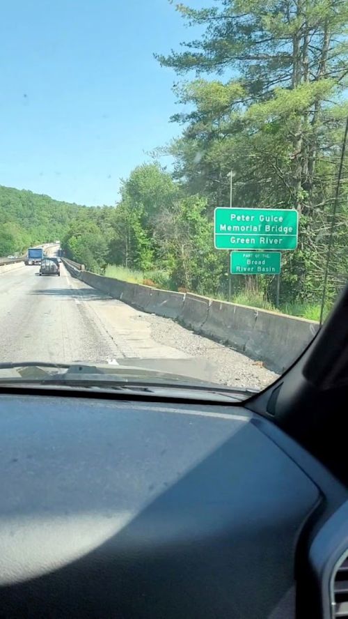 View from a vehicle showing a highway sign for Peter Gulce Memorial Bridge over the Green River, surrounded by trees.
