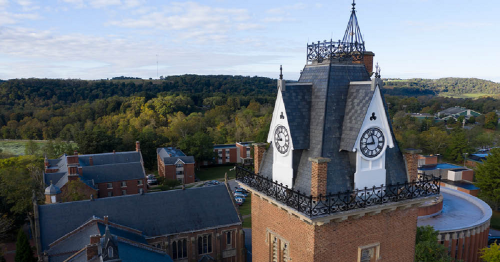 Aerial view of a historic building with a clock tower, surrounded by trees and other structures in a scenic landscape.