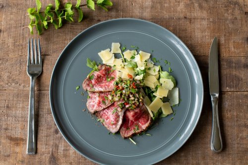 A plate of sliced beef with herbs, cheese shavings, and a fresh salad, set on a rustic wooden table.